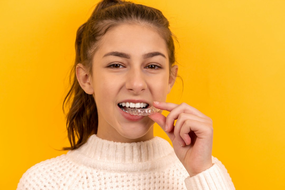 Smiling young woman on a yellow background placing a clear removable appliance on her teeth